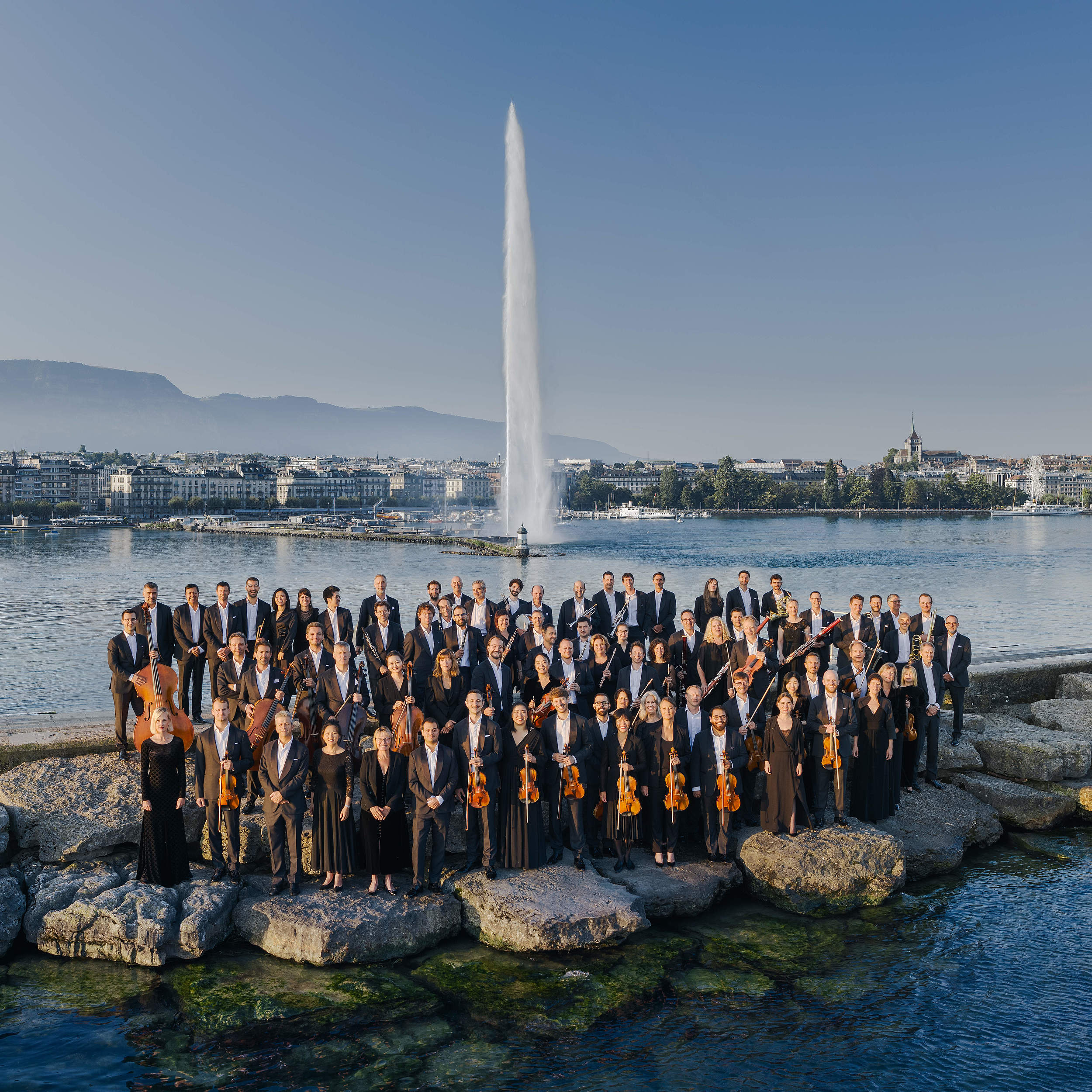 Photo de l'Orchestre de la Suisse Romande posant devant le Jet d'Eau de Genève, emblématique monument suisse, associant l'élégance de la musique classique à la beauté du paysage genevois.