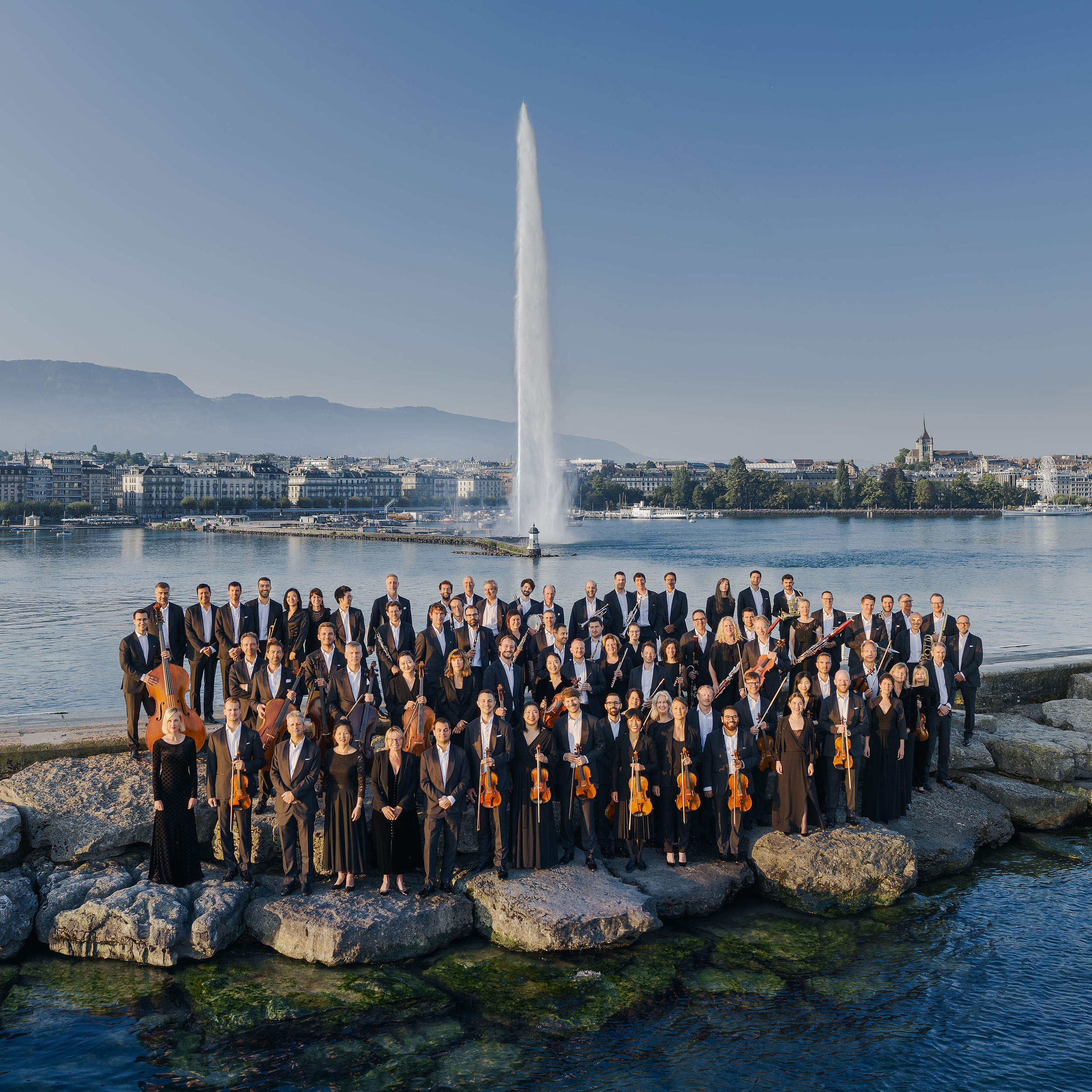 Photo de l'Orchestre de la Suisse Romande posant devant le Jet d'Eau de Genève, emblématique monument suisse, associant l'élégance de la musique classique à la beauté du paysage genevois.