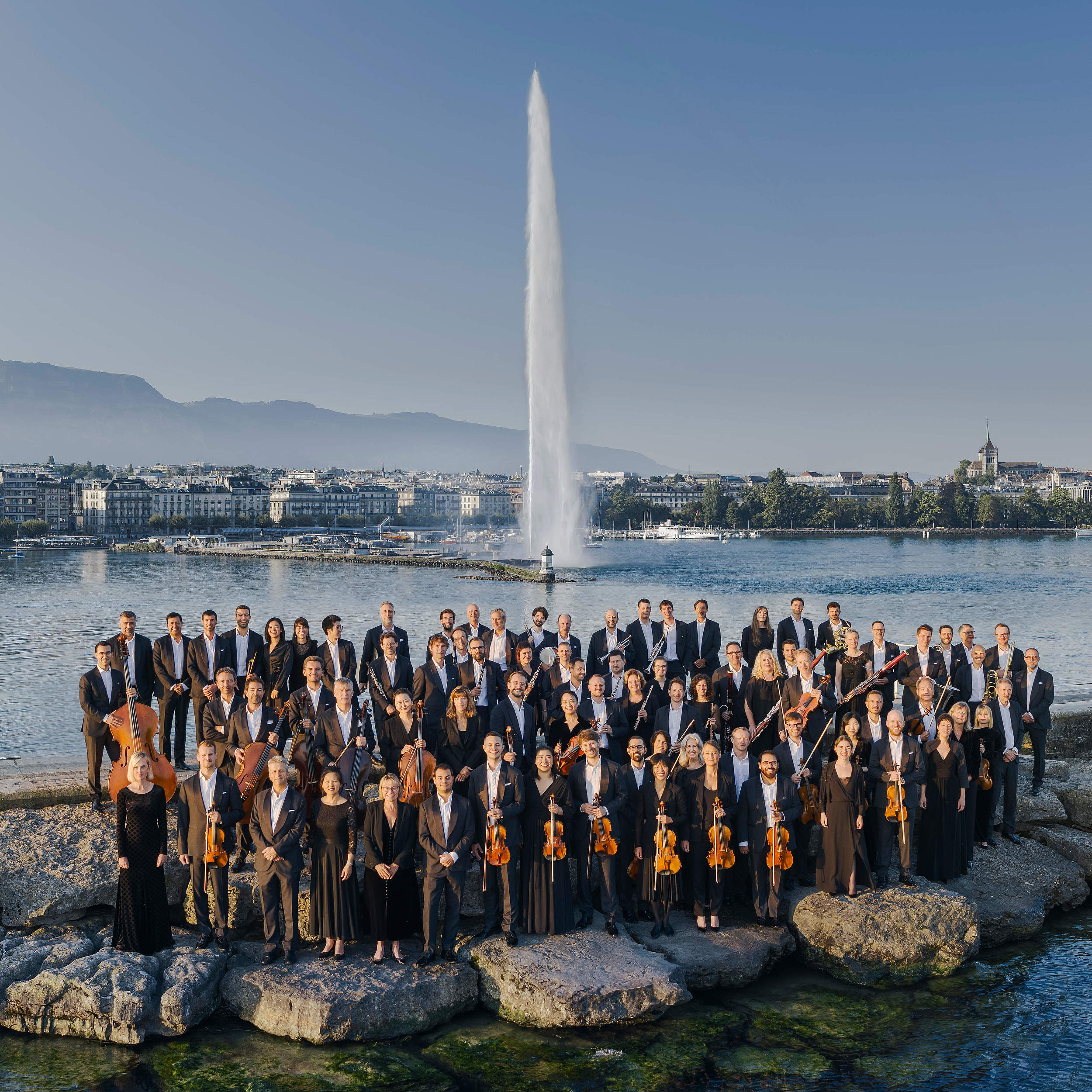 Photo of the Orchestre de la Suisse Romande posing in front of Geneva's Jet d'Eau, an iconic Swiss monument that combines the elegance of classical music with the beauty of the Geneva landscape.